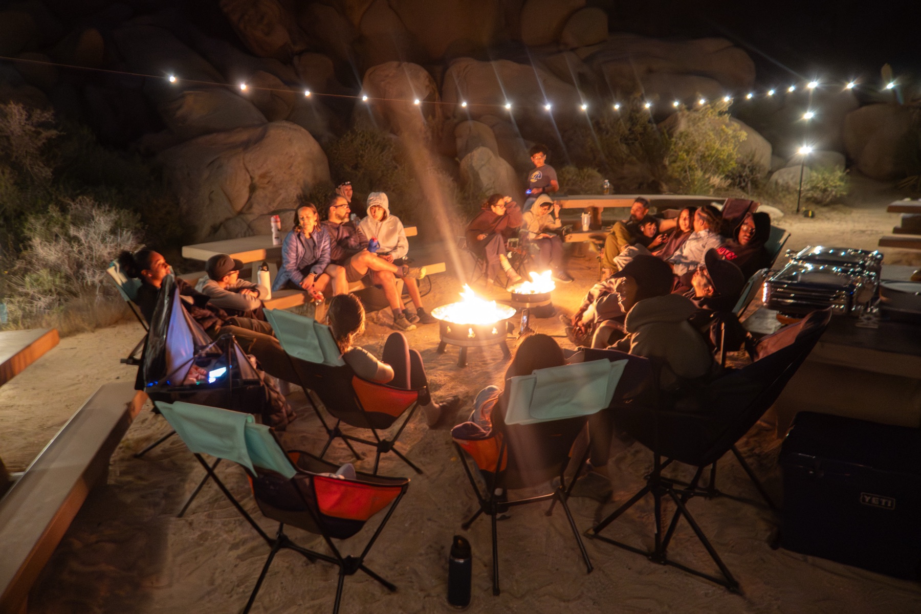 Families gathered in camp chairs around a campfire under string lights after dark